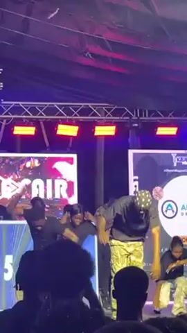 A man is performing on stage at an event with two dancers. He is wearing a black t-shirt, camouflage pants, a beaded necklace and a black and white bandana on his head. He is standing up and dancing, moving his body to the beat of the music. The stage is lit with multiple colored lights and there are two large screens on either side of the stage. The screen on the right has a logo for a company called AnyCar Deals GH. The event appears to be sponsored.