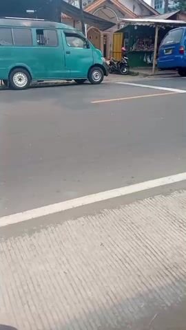 A green van is parked on the left side of the road. A motorcycle is parked behind the van. A person wearing a helmet and a backpack is riding a red motorcycle in the middle of the road. Another person is riding a blue and white motorcycle in the middle of the road. A blue truck is parked on the right side of the road. There is a small market stall on the right side of the road. The road has white lines painted on it. The weather appears to be sunny.