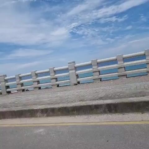The video shows a view from a moving vehicle, likely a car, traveling along a road next to a body of water. The road surface is visible in the foreground, with a concrete curb and a sidewalk beyond it. A white, segmented railing runs along the sidewalk, separating it from the water. The water appears to be a bright blue, with some whitecaps. The sky is a clear blue with scattered white clouds. In the distance, on the left side of the frame, a white tower with a circular balcony is visible, surrounded by a metal fence with barbed wire. A utility pole with wires is also visible. The vegetation along the roadside consists of green bushes and some taller trees. The overall scene suggests a coastal location on a sunny day.