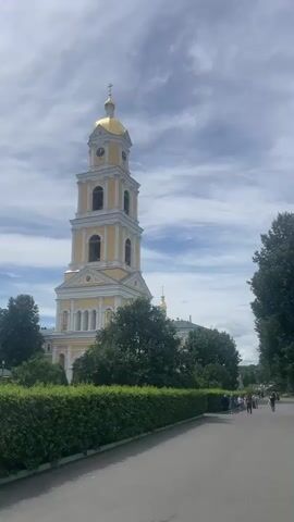 The video shows a tall, yellow bell tower with a golden dome against a cloudy blue sky. A path runs in front of a neatly trimmed green hedge. In the background, people are walking along the path, and trees are visible.