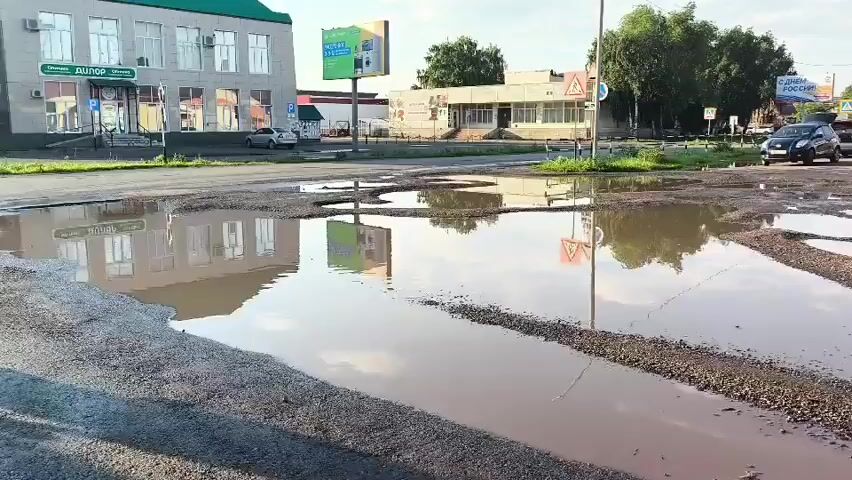 The video shows a street with numerous large potholes filled with muddy water. Cars are visible driving on the street, navigating around the potholes. Buildings and trees are seen in the background. The sky is bright, suggesting it is daytime. The overall condition of the road appears to be poor.