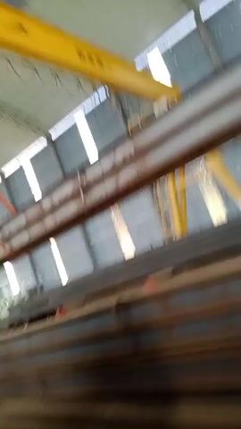 The video shows a large industrial warehouse with a high ceiling. A yellow overhead crane is moving a long steel beam. The beam is suspended from the crane by orange straps. In the background, there are stacks of other steel beams. There are at least two people visible, wearing orange vests, who appear to be workers. The lighting is artificial, suggesting an indoor environment. The overall impression is one of industrial activity and material handling.