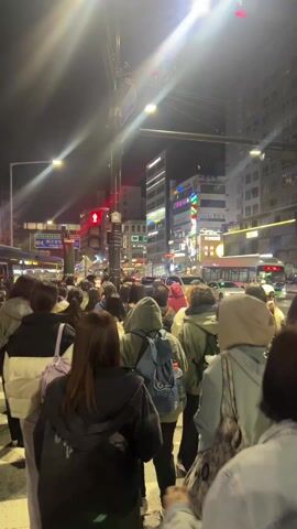 A nighttime scene shows a crowd of people crossing a zebra-striped crosswalk in a city. The view is from within the crowd, looking towards buildings lit up with signs and streetlights. A green and white bus drives by on the right. A police officer wearing a high-visibility yellow vest and a mask stands near the crosswalk, observing the pedestrians. Several cars are seen driving in the distance. The crosswalk light is red, but the crowd continues to cross.