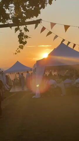 The video depicts an outdoor evening event or celebration at sunset. Multiple white party tents are set up on a grassy area, with guests seated at tables inside the tents. A white dance floor is positioned between two tents. In the background, the sun is setting, casting an orange glow across the sky and distant hills. Colorful triangular bunting is strung above the tents. In the foreground, a vine-covered pergola or archway is visible on the left, and a few individuals are standing, one possibly filming. People are gathered, some seated and others moving around, suggesting a social gathering like a wedding reception.