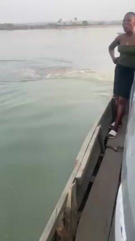 A wooden boat is moving on a body of water that appears to be a river or a lake. The water's surface is active, with ripples and a muddy patch visible beneath. On the boat, a person wearing white sandals stands by the side. The camera's angle shifts, and the focus is directed to the river's surface. Later, the camera's focus shifts again to capture a woman standing on the boat, wearing a green top and black skirt, positioned near a silver car. Behind her, other individuals can be seen in the boat, along with additional boats in the distance.