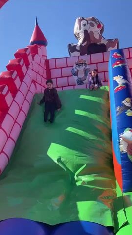 The video shows a young girl going down a green inflatable slide on a sunny day.  The slide is attached to a large inflatable castle with red and pink brick designs. At the top of the slide, another girl is sitting near cartoon decorations of Chip and Dale characters. A third girl stands at the bottom of the slide, observing. The inflatable structure is blue at its base and features a colorful design on the side near the ladder.