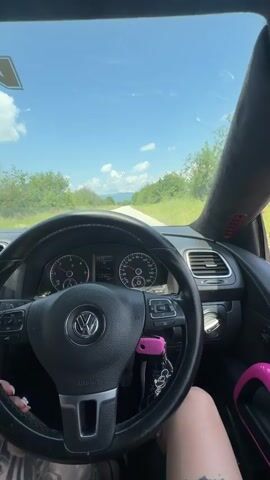 The video captures a first-person perspective from inside a car, likely a Volkswagen, driving on a sunny day in Krasnovo, Bulgaria. The driver's hands, adorned with tattoos and light-colored nails, rest on the black leather steering wheel, which prominently features the Volkswagen logo. A bright pink car key, with multiple keychains attached, is inserted into the ignition on the right side of the steering column. Matching pink accents are visible on the door trim and a panel on the A-pillar.

Through the steering wheel, the dashboard gauges are visible, showing a low speed (between 10-20 units, likely km/h) and an odometer reading of approximately 173,204. The view through the windshield reveals a clear blue sky with scattered white clouds. A paved road stretches ahead, flanked by lush green trees and bushes. In the distance, rolling hills or mountains are visible under the bright sunlight. A tattoo is also visible on the driver's right leg. The scene depicts a serene drive through a rural or scenic landscape.