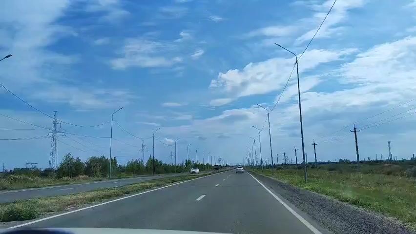 A first-person perspective video shows a car driving down a two-lane highway on a clear day. The sky is blue with some white clouds. There are streetlights and power lines on either side of the road. Trees and bushes line the left side of the highway, while a grassy field is on the right. Several cars and a truck can be seen on the road ahead. The car appears to be in motion.