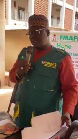 Here's a description of the video:

The video shows a man speaking into a microphone while holding a stack of papers. He's wearing glasses, a patterned hat, an orange shirt, and a green vest. The vest has "FMAFS" and "FEDERAL MINISTRY OF AGRICULTURE AND FOOD SECURITY" printed on it. There is a Nigerian flag on the vest as well.

He stands behind a table with a book on it. Behind him, a banner displays "FMAFS FEDERAL MINISTRY OF AGRICULTURE AND FOOD SECURITY HOPE." The background also includes a multi-story building. The video is taken outdoors during the daytime.