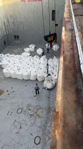 The video shows a large cargo ship's hold being loaded with large white sacks. A crane is visible, lifting several sacks at once. Three workers in hard hats are present in the hold, directing the loading process. One worker stands near a pile of sacks, another is on top of a pile of sacks, and a third is near the crane. The hold is cavernous and appears to be made of metal. A "NO SMOKING" sign is visible on the far wall. The environment suggests an industrial port or shipping facility.