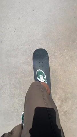 The video opens with a top-down view of a person's feet on a black skateboard. The individual wears green and white sneakers with white socks and brown cargo shorts, with the sun casting a shadow directly behind their legs. They briefly push off the ground to initiate movement.

The camera then pans upwards and forwards, revealing a concrete path on what appears to be a bridge. A sturdy metal railing runs along the right side of the path. Below the bridge is a wide, verdant landscape filled with dense green vegetation, stretching into the distance under a clear, bright blue sky. Power lines and large power pylons are visible against the skyline, with distant buildings and hills completing the horizon.

The scene shifts to a wider perspective, showing two individuals from behind as they move along the bridge path. The person from the initial shot, still in green sneakers, is skateboarding. Alongside them is another person, also in shorts and a dark top, carrying a backpack. A dark car passes on the road adjacent to the path. The sun is high and bright, indicating a clear, warm day, and casting long shadows from the railing and the people onto the concrete. The setting suggests an outdoor, possibly semi-urban or park-like environment, on a bridge in Santa Perpètua de Mogoda, Spain.