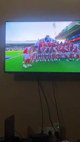 A person is filming a television screen showing a soccer team, dressed in red and white uniforms, celebrating their victory on a green field. They are gathered around a trophy. The camera angle is looking slightly up at the TV screen, which is mounted on a beige wall.  A laptop and an electronic device with cables are visible on a surface below the television. The video is likely taken indoors.