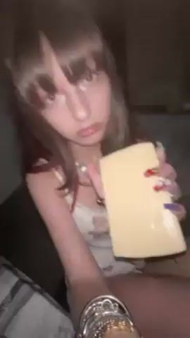 A young woman with long brown hair and bangs is seated and eating a large block of yellow cheese. She is wearing a white tank top with a teddy bear print, multiple necklaces, and bracelets. Her fingernails are long and decorated with colorful designs. She takes a bite of the cheese, looks at the camera, and smiles. The background is dark, suggesting an indoor setting, possibly a car.