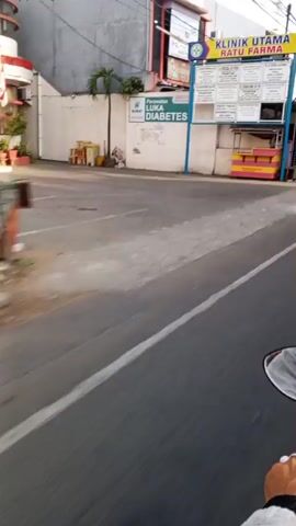 The video begins with a view of a street with a gray concrete sidewalk and an asphalt road. A building with white walls and a blue trim is visible on the left, featuring signs that read "LUKA DIABETES" and other unreadable text. Further down, a yellow and pink food stall with a white canopy is situated. The camera then pans to the right, showing a person wearing a red hijab and a gray shirt riding a yellow bicycle with yellow wheels. The person is carrying a sign on the handlebars. Behind them, there is a multi-story building with a black and white facade. The video continues down the street, revealing more buildings, storefronts with closed blue and green doors, and parked vehicles, including a yellow becak (a three-wheeled bicycle taxi). The camera captures glimpses of various signs and posters on the buildings, some with images of people. The environment appears to be an urban or semi-urban setting, possibly in Southeast Asia, with a mix of residential and commercial properties. The lighting suggests it might be daytime, with some areas in shadow. The video concludes with a shot of the street from a moving perspective, showing more of the surrounding buildings and infrastructure.