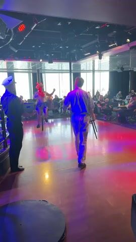 The video captures a lively indoor performance. Three individuals are the main subjects on a stage bathed in vibrant red and purple lighting. In the foreground, a person in a dark uniform with a white hat stands facing away from the camera, observing the scene. Center stage, a performer in a blue costume, possibly a jester or clown with a distinctive headdress, is skillfully balancing on a unicycle. To the right of the unicyclist, another person, dressed in a striped shirt and white pants, is interacting with the performer.

The scene appears to be a performance venue, likely a club or a theater, with an audience seated in the background. Large windows behind the stage offer a glimpse of an outdoor environment, suggesting it could be daytime or twilight. The audience, composed of various individuals, seems engaged and entertained by the performance. The atmosphere is energetic and festive, amplified by the colorful stage lighting. The overall impression is that of a comedic or variety show in progress. There is no visible text in the frame. The location context of Espoo, Finland, does not provide specific cues within the image itself.