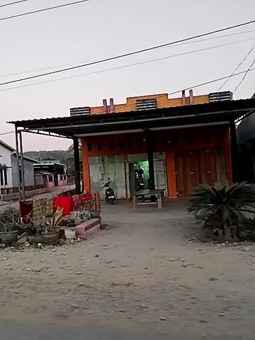 The video shows a street scene in what appears to be a rural or semi-rural area. The street is unpaved and dusty, with some potholes visible. On either side of the street are small buildings, some of which appear to be houses and others shops. There are also some trees and vegetation. The sky is overcast, suggesting it is either early morning or late afternoon. The video pans from left to right, showing different sections of the street and the buildings along it. In the foreground, there are some potted plants and other miscellaneous items placed outside one of the shops. One building, in particular, is painted orange and has roll-up doors, suggesting it is a commercial establishment. Another building is a taller, multi-story structure that appears to be made of concrete blocks. The overall impression is of a quiet, somewhat undeveloped area.