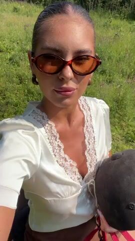A young woman with her hair pulled back and wearing sunglasses and a white top with lace trim sits on a horse. She is holding a black riding helmet. The background is a grassy field with trees in the distance. She turns her head and smiles at the camera.