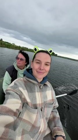 Two women are in an inflatable boat on a lake. The woman in the foreground is wearing a plaid jacket and a headband with frog eyes. She is smiling and holding a phone to take a selfie. The woman in the background is wearing sunglasses and a green hoodie. The sky is cloudy and the water is calm with small ripples. The boat is moving through the water, indicated by the wake behind it. The shoreline with trees and vegetation is visible in the distance.