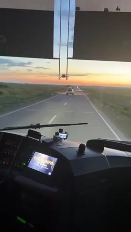 This media file captures the view from the driver's seat of a vehicle, likely a bus or truck, as it travels down a highway in Kazakhstan. The scene is set during a sunset, with the sky painted in hues of orange, yellow, and blue. The sun is low on the horizon, casting a warm glow over the landscape.

The road ahead stretches into the distance, with a single car visible in front, its brake lights illuminated. The surrounding environment is rural, characterized by vast, open fields of green grass that extend to the horizon. The weather appears clear and calm, conducive to travel.

Inside the vehicle, the dashboard is visible, featuring a multimedia screen displaying the title "СЕДАЯ НОЧЬ" (Sedaya Noch), which translates to "Gray Night." This suggests music is playing, contributing to the atmosphere of the journey. The overall emotion evoked is one of peaceful solitude and the open road, with a sense of journeying towards or away from the setting sun. The time of day is clearly evening, as indicated by the sunset.