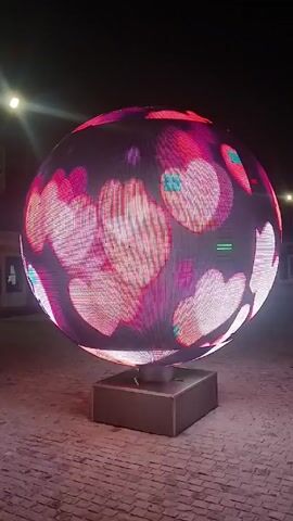 A spherical LED screen displaying pink hearts sits on a square base at night. The screen is pixelated and the background is dark with some streetlights and buildings visible. The camera remains stationary.