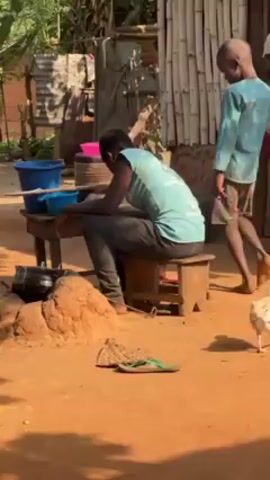 In a rural outdoor setting, a young man wearing a light blue tank top and grey pants sits slumped over on a small wooden stool. His head is bowed, and he appears to be exhausted. Behind him, a slightly younger child with a white knit hat pulls playfully at his shirt. To the left, an even younger child in a blue shirt stands partially visible. The man sits near a makeshift outdoor kitchen area; a pot sits on a clay-built stove. A wooden table holds various colorful wash basins.  A chicken walks in the foreground near discarded green flip-flops. The ground is red dirt, and sunlight dapples the scene.  A cluttered array of wooden structures, corrugated metal sheets, and foliage form the backdrop.