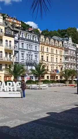 The video shows a street scene with people and buildings. In the foreground, there is a large white sign that says "I LOVE KARY". Two people are standing near the sign. In the background, there are several multi-story buildings with elaborate architecture. Some of the buildings have balconies and many windows. Palm trees are visible on the left and right sides of the sign. The sky is blue with some white clouds. The ground is paved with cobblestones. In the middle ground, other people are walking around and interacting. Some benches are visible. One building in the background has a sign with a swan logo and the word "Bata". The scene appears to be a tourist area, possibly a town square.