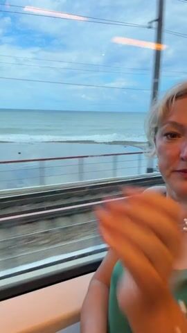 A woman with blonde hair is sitting on a train, looking out the window. She is wearing a green dress and pearl jewelry. Outside the window, the ocean and beach are visible. The train tracks are also visible in the foreground. The sky is cloudy. The camera is focused on the woman's face and profile, and also shows her hand resting on her shoulder.