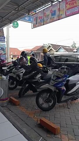 The scene takes place under the awning of an Alfamart convenience store in what appears to be a residential area. Several motorbikes, including Honda models, are parked closely together on a paved surface marked with yellow lines and short orange bricks. The sky is overcast, giving the scene a somewhat muted quality. A woman wearing a light pink top and a hijab carries a young child. She walks past a parked black car, towards the side of the building. Various advertisement posters are displayed beneath the awning, promoting different products. The backdrop includes single-story houses with visible roofs. The atmosphere seems ordinary and casual.