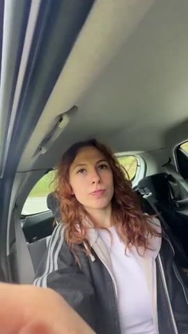 The video begins with a close-up of a woman with reddish-brown wavy hair sitting in a car, winking. She is wearing a white shirt and a grey and black striped jacket. The camera then pans up, showing trees and a blue sky with clouds through the car window. The camera continues to move upwards, revealing a scenic view of mountains, green trees, and a winding road. The car is white, and the door is open.