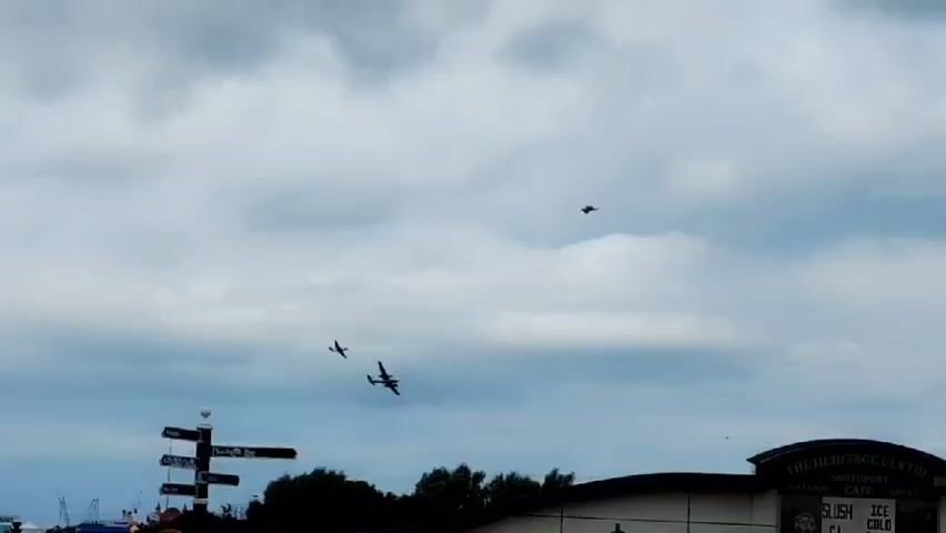 The video shows three airplanes flying in formation against a cloudy sky. The planes appear to be vintage aircraft, possibly from the World War II era. One plane is in the foreground, while the other two are further back. The planes are flying over a building that has the words "THE HERITAGE CENTRE" visible on it. There are also trees and a lamppost in the foreground. The overall scene appears to be a public event or airshow.