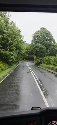 The video shows a view from inside a vehicle as it drives on a wet, winding road lined with lush green trees. The road has white double lines and what appear to be rumble strips. There is a yellow and black chevron road sign visible on the left side of the road, indicating a sharp curve. The sky is overcast.