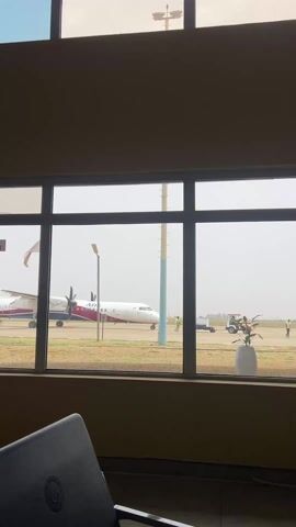 The video shows an Arik Air turboprop plane on a runway, viewed through a window. The plane is white with maroon and dark blue accents, and its propellers are visible. There is a small Nigerian flag on the plane near the front. A streetlight and other poles are visible on the tarmac along with a utility vehicle and a piece of ground equipment. A person in a yellow vest walks in the distance. Inside, part of a chair and a potted plant are visible in the foreground. The light is hazy.