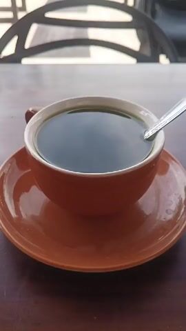 A close-up, static shot of a dark liquid in an orange ceramic mug with a silver spoon resting on the rim. The mug sits on an orange saucer, which is on a wooden table. In the background, a blurred chair with a patterned seat is visible. The lighting is natural, suggesting an outdoor or well-lit indoor setting. There are no people or other significant objects in the frame. The video is extremely short, showing no discernible action.