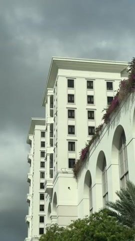 The media file captures a close-up, low-angle view of a modern, white building against a dramatic, overcast sky. The building features a tiered design with multiple stories, each adorned with evenly spaced dark-framed windows. Ornate architectural details, such as balconies with pillars and arched windows on a lower section of the building, are visible. A lush display of purple flowers cascades down from a terrace on one side of the building, adding a touch of vibrant color. Green foliage from trees peeks in from the bottom of the frame. The overall impression is one of contemporary elegance and a sense of impending or recent rain due to the dark, heavy clouds. The time of day appears to be late afternoon or early evening, given the low light. No people are visible, and the scene focuses entirely on the architectural grandeur of the building and the atmospheric weather. The location context provided suggests this building is situated in Coral Gables, Florida, in the United States. There is no visible text in the media file.