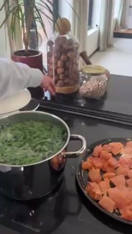 On a black cooktop, a pot of green beans is boiling, and next to it is a frying pan full of cubed raw chicken. Behind the cooktop, on a black counter, is a jar filled with brown objects, and a smaller jar filled with white objects. Behind the counter is a large houseplant and a window. A hand reaches into the frame, and moves a white plate to the left side of the frame. The cooktop is labeled "Electrolux". A small "Siemens" label is visible on the wall between the cooktop and the cabinet above.