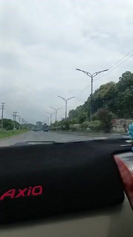 A video taken from the interior of a car shows a highway scene. The camera is positioned looking forward through the windshield. A black dashboard mat with the red text "AXIO" is visible in the foreground. Outside the car, the highway stretches ahead, lined with trees and streetlights on both sides. Several vehicles, including cars and a bus, are driving on the road. The sky is overcast with gray clouds. The video appears to be a first-person perspective of driving on a road.