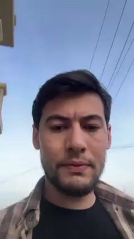 A man films himself walking along a street. He is wearing a brown and white flannel shirt over a black shirt. He has short brown hair and a short beard.  Behind him, the background consists of a street with cars, sidewalks, buildings, and trees. The sky is overcast.