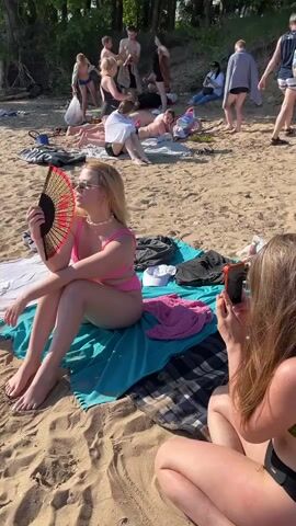 The video shows a beach scene with several people. In the foreground, a young woman with blonde hair wearing a pink bikini is sitting on a teal towel and fanning herself with a red and black fan. Next to her, another young woman with long brown hair, wearing a green and black bikini, is kneeling and taking a photo of the first woman with her phone. Behind them, several other people are scattered on the sand, some lying down, some sitting, and some standing. The environment appears to be a sunny day at a beach with sand and vegetation in the background.