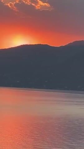 The video shows a sunset over a body of water. The sun is setting behind a dark, silhouetted mountain range. The sky is a vibrant orange and red, with some clouds visible. The water in the foreground reflects the colors of the sunset, creating a warm and peaceful atmosphere. There are no people or man-made objects visible in the scene.