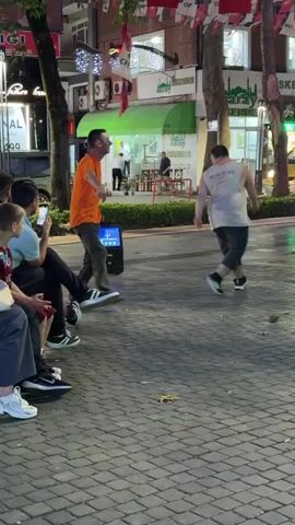 Two men are dancing in the street. One man is wearing an orange shirt and dark pants. The other man is wearing a light-colored tank top and dark shorts. They are both facing away from the camera, but their bodies are angled towards each other. There is a portable speaker on the ground between them. Several people are sitting on the side of the street, watching the dancers. The street is paved with cobblestones, and there are trees and buildings in the background. Turkish flags are hanging from wires strung across the street. It appears to be nighttime, and there are lights from the buildings and streetlights illuminating the scene.