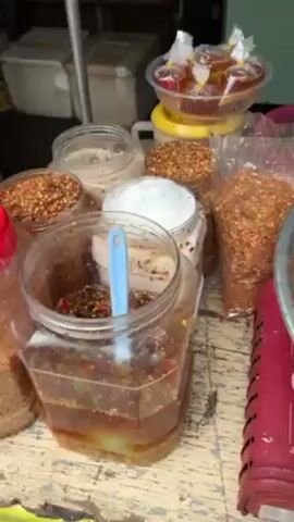 The video shows a person preparing a dish outdoors. The person is wearing gloves and working behind a table laden with ingredients and condiments.  In front of the person is a large metal bowl filled with what appears to be small, colorful shellfish mixed with green herbs and pieces of red chili pepper. A tin can is partially submerged within the mixture.  Nearby, there are jars and bags containing various spices, seasonings, and sauces. Limes are visible in a green bowl. The table itself is slightly worn and appears to be set up on a portable gas stove.