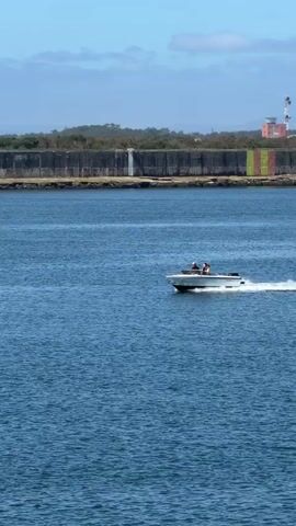A white speed boat with two occupants is traveling across a blue body of water, leaving a white wake. In the background, there is a long, concrete structure running along the shoreline, with some vegetation on top of it. Above the structure, the sky is partly cloudy. Further in the distance, a white cylindrical tower is visible.