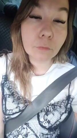 The video begins with a close-up of a woman's face as she is in a car, wearing a white t-shirt over a black and white lace camisole. She has long brown hair, false eyelashes, and is wearing a seatbelt. The background is blurred but appears to be the interior of a car. The woman speaks and smiles at the camera. The footage continues with slight variations in her expression and head movements, but she remains seated in the car. The lighting suggests it is daytime, possibly overcast.