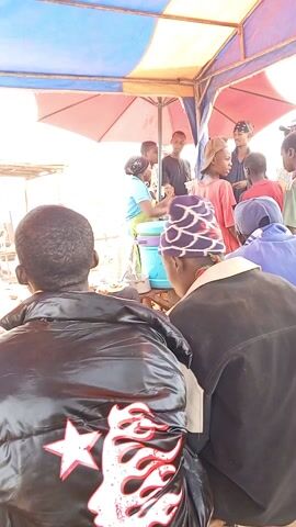 The video shows a busy outdoor food stall scene. Several people are standing near the stall, which is covered by a blue and red tent. A woman in a blue shirt and green apron is serving food from a blue container to customers. Some customers are sitting down, their backs to the camera, while others are standing and waiting for their orders. The stall is surrounded by other temporary structures, possibly other stalls or seating areas. The overall atmosphere suggests a lively and communal eating experience.