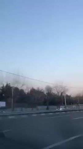 The video, shot from inside a moving vehicle, shows a road with a concrete barrier on one side. Beyond the barrier are trees, and in the distance, a plume of smoke is visible in the sky. Street lights line the road. The sky is clear and blue.