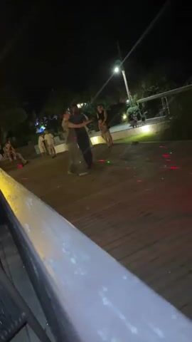 A nighttime outdoor scene shows a wooden deck with people dancing and moving. Green and red laser lights are projected onto the deck, creating patterns. In the center, two individuals are dancing, one in a dark shirt and the other in a lighter gray shirt. Several other people are visible in the background, some sitting and some standing. The environment appears to be an entertainment area with lighting and foliage.