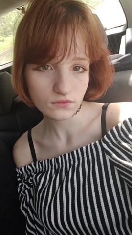 A young woman with short red hair and a septum piercing is sitting in a car. She is wearing a black and white striped off-the-shoulder top and a black choker. She looks at the camera and smiles slightly. The background shows the car's interior and a glimpse of green trees outside.