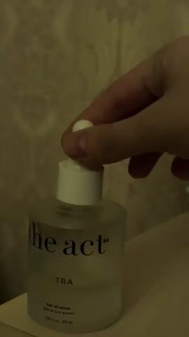 A hand is seen squeezing a white dropper, releasing a drop of clear liquid into the opening of a clear glass bottle. The bottle has black text that reads "the act" and "EXTRA" along with smaller text indicating it is hair oil serum. The background consists of a patterned wallpaper and a light-colored surface.