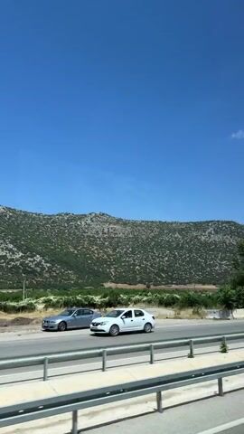 The video shows a view from a vehicle moving on a road. The road has a guardrail on the right side of the frame. A white car is seen driving on the road. In the background, there is a large, green, and rocky mountain under a clear blue sky. Trees are scattered along the side of the road and in front of the mountain. There is a small, dilapidated structure with a white roof and white walls near the left side of the frame, further back from the road. The overall scene depicts a rural or mountainous landscape.