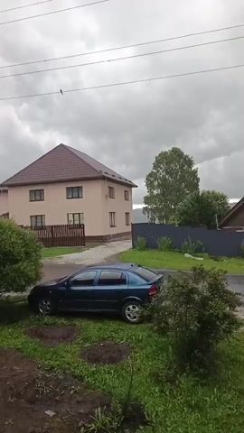 This media file captures a serene outdoor scene with a focus on houses and vehicles under a cloudy sky. The setting appears to be a residential area, possibly a village or suburb, given the presence of houses, fences, and parked cars.

In the foreground, a blue hatchback car is parked on a patch of grass near a leafy bush. To the right, the front of a black car is visible, partially obscuring the view of the blue car. In the background, two houses are prominent. The larger house on the left is a light-colored, multi-story building with a brown roof. A wooden fence separates it from the street. The house on the right is smaller, with a dark roof and white-framed windows, set behind a dark grey fence. A tall, slender tree stands between the two houses, and more greenery and trees are scattered throughout the scene. The ground in front of the blue car is muddy, suggesting recent rain.

The weather appears to be overcast, with a thick layer of grey clouds obscuring the sun. This creates a somewhat somber or tranquil atmosphere. The overall impression is one of a quiet, perhaps rural or semi-rural, residential environment on a cloudy day, likely in the afternoon or evening, given the diffused light. No people are visible, and no text is present in the video. The scene conveys a sense of everyday life in a seemingly peaceful setting.